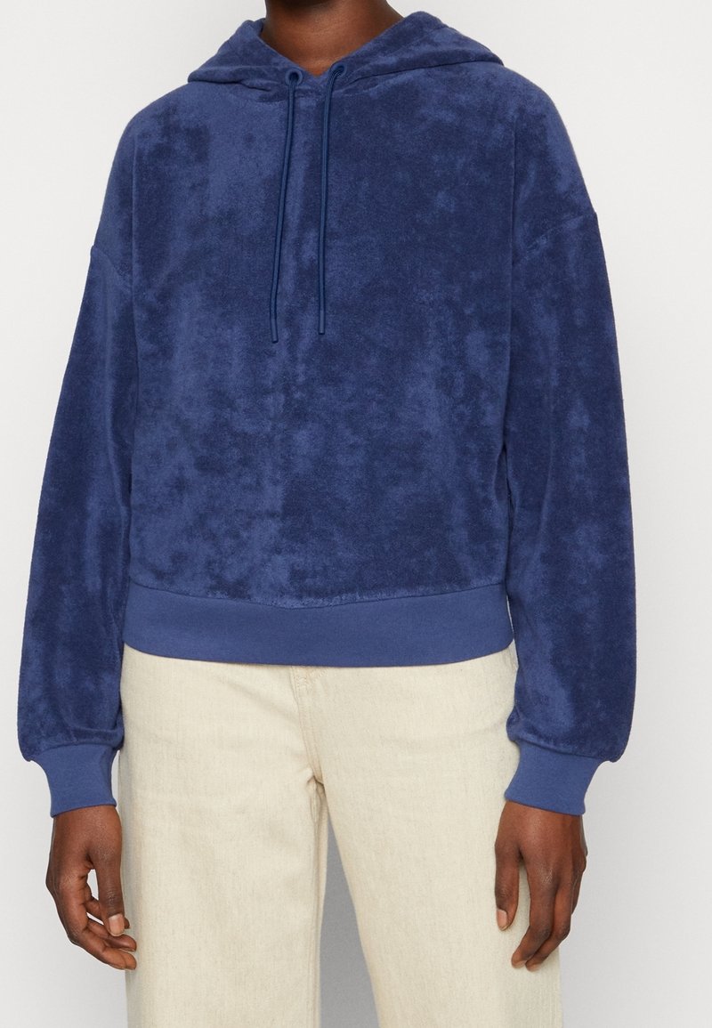 Granatowy polarowy sweatshirt z kapturem, z sznurkiem do regulacji, ściągaczami przy mankietach i na dole, zaprezentowany na tle jasnych spodni.