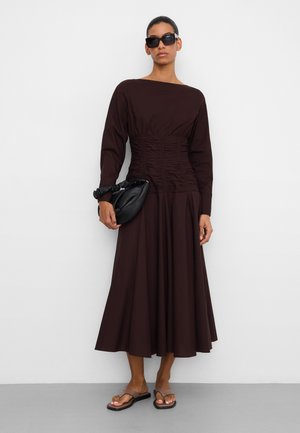 Kobieta w ciemnobrązowej, marszczonej sukience midi z długim rękawem, czarnych okularach przeciwsłonecznych, trzymająca czarną, marszczoną kopertówkę i nosząca czarne płaskie sandały.