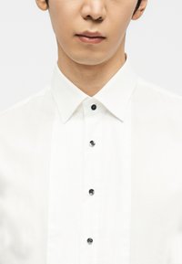 Εγγύς λήψη ενός ατόμου που φορά λευκό πουκάμισο με πιέτες στο μπροστινό μέρος και μαύρα κουμπιά, χωρίς να είναι ορατά χαρακτηριστικά του προσώπου πάνω από τη μύτη.