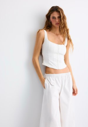 Kobieta z długimi falowanymi włosami ubrana w biały dopasowany krótki top i luźne białe spodnie, stojąca na tle jednolitej jasnej ściany.