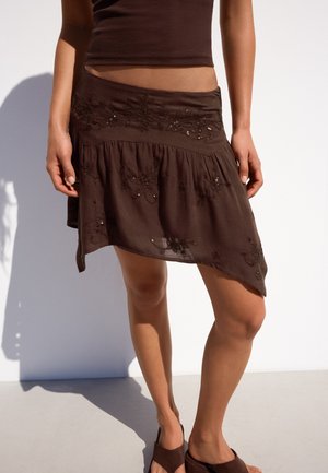 Femme portant une jupe asymétrique marron avec des détails floraux brodés, assortie à un top court assorti et des sandales.