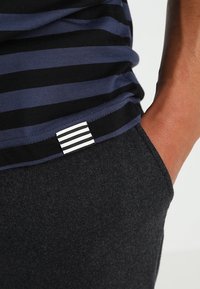 Camisa a rayas en azul marino y negro con una etiqueta a rayas en blanco y negro, combinada con pantalones de tela gris oscuro, que muestran una textura suave y un corte ajustado.