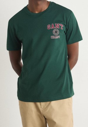 Man wearing a dark green GANT Champs t-shirt with short sleeves and beige pants, standing with hands behind back.