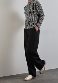 Top a maniche lunghe bianco e nero con un motivo geometrico, abbinato a pantaloni neri e sneakers bianche, in piedi su una superficie grigia.