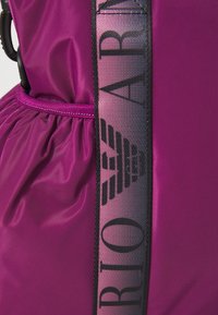 Lilla nylonveske med en samlet kropp og en kontrastrem som har svart tekst og et logomønster. Glatt tekstur, funksjonell design.