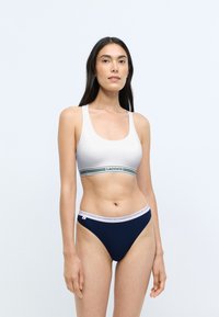 Witte katoenen bralette met een groene en marineblauwe gestreepte band met het "LACOSTE" logo; gecombineerd met marineblauwe low-rise bikini slipjes met een witte tailleband.