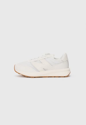 Zapatilla deportiva blanca con suela de goma, parte superior de malla y cuero, cierre de cordones y un gran logo "N" en el lateral, mostrada de perfil.