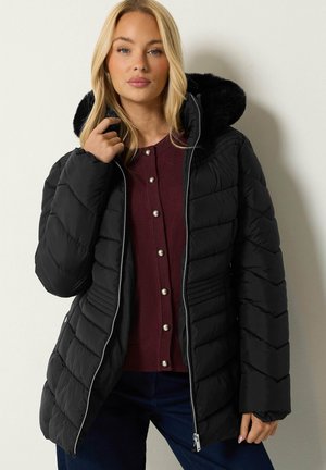 Femme portant un manteau d'hiver matelassé noir avec capuche bordée de fourrure et un pull bordeaux boutonné, debout sur un fond clair.