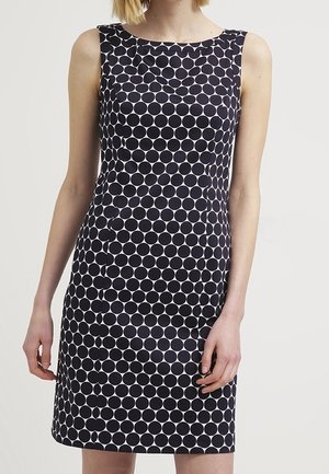 Ärmelloses, figurbetontes Kleid mit großem schwarz-weißem Polka-Dot-Muster, getragen von einer Person, die vor einem einfarbigen hellen Hintergrund steht.