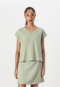 Top verde chiaro a maniche corte con scollo a V, design a strati. Il tessuto sembra leggero e ha una morbida texture; indossato sopra una gonna abbinata.