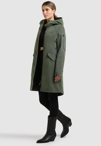 Femme portant un manteau à capuche vert olive, un crop top noir, des leggings noirs et des bottes en cuir noires mi-mollet, debout avec les jambes croisées.