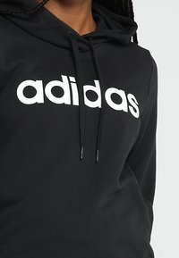Sudadera negra de tela suave con un gran logo blanco de "adidas" en el frente. Incluye cordones en la capucha.