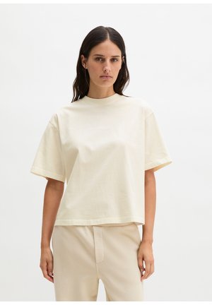 Kobieta o długich, ciemnych włosach, ubrana w prostą, jasnobeżową, oversize koszulkę z krótkim rękawem i pasujące spodnie na białym tle.