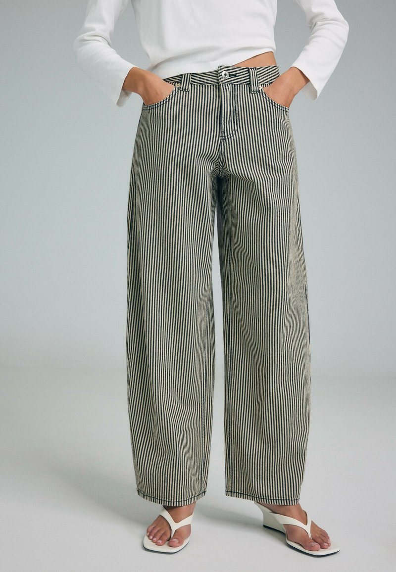 Personne portant un pantalon large taille haute à rayures verticales noires et blanches, les mains dans les poches et des sandales blanches à talons.