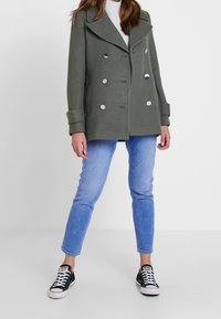 Abrigo de color verde oliva con doble botonadura, grandes solapas y botones plateados. Llevado sobre un suéter blanco y jeans azules, combinado con zapatillas negras.