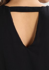 Musta yläosa, jossa on syvä V-muotoilu selässä. Valmistettu teksturoidusta kankaasta, jossa on ohut kaula-aukko, luoden minimalistisen ja elegantin siluetin.