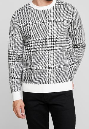 Mężczyzna ubrany w sweter w czarno-biały wzór pepitki z białymi mankietami i krawędzią, do tego czarne spodnie na szarym tle.