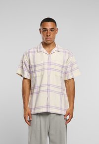 Camicia a maniche corte a quadri color crema con strisce viola e lavanda, dotata di due tasche sul petto e una vestibilità rilassata. Materiale in cotone testurizzato.