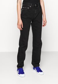 Jeans de mezclilla negra con corte recto, que cuentan con bolsillos frontales, botones de metal y un ligero desgaste. Se usan con zapatillas deportivas azules.