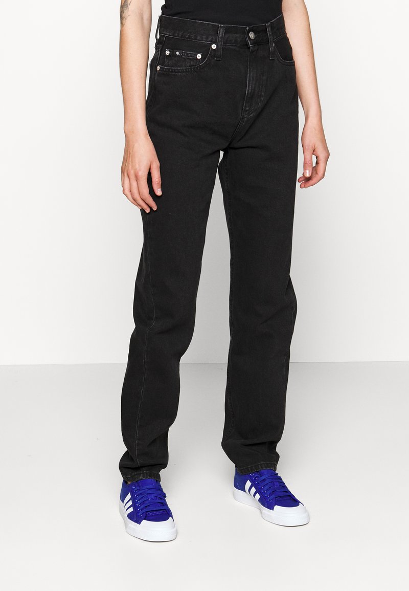 Jeans de mezclilla negra con corte recto, que cuentan con bolsillos frontales, botones de metal y un ligero desgaste. Se usan con zapatillas deportivas azules.