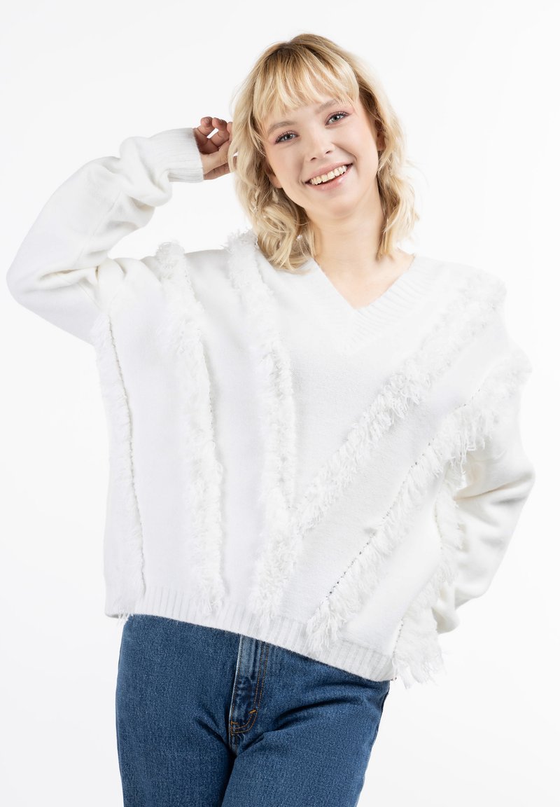 Biały, oversize'owy sweter z dekoltem w serek, z teksturowanym frędzlowaniem ułożonym w diagonalne linie. Noszony z niebieskimi dżinsami.