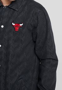 Mörkgrå jacka med broderad röd och vit tjur-logotyp på bröstet och en diskret upprepande textmönster över tyget.