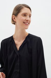 Černá košile s knoflíky vyrobená z měkké, mírně texturované tkaniny. Má nařasený výstřih a dlouhé rukávy, což vytváří uvolněný střih.