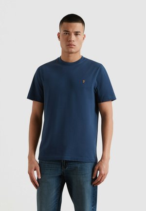Mężczyzna ubrany w gładki granatowy t-shirt z krótkim rękawem i niebieskie dżinsy, stoi z ramionami rozluźnionymi na tle jednolitego jasnego tła.