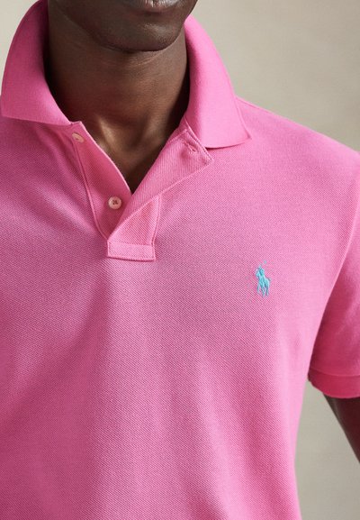 Ροζ πόλο πουκάμισο από υφασμάτινη υφή, με γιακά και μπλε κεντητό λογότυπο αλόγου στο αριστερό μέρος του στήθους.