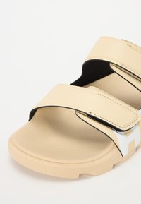 Beige sandaalit, joissa on kaksi leveää hihnaa, sileä pinta, tarranauha-kiinnitys ja paksu pohja, jossa on kuvioitu alaosa ja valkoinen logo.