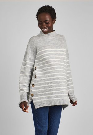 Smilende kvinde iført en løs grå sweater med hvide vandrette striber og knapper i siden, kombineret med blå jeans.
