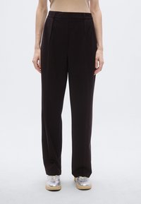 Pantalon à jambes larges en tissu brun foncé ; présente des plis devant, une coupe ajustée, des poches latérales et une texture lisse. Associé à des baskets argentées.