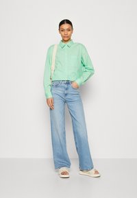 Camisa de botón verde claro con un bolsillo en el pecho, combinada con pantalones vaqueros anchos de cintura alta en azul claro y sandalias de plataforma color crema.