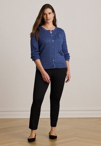 Femme debout à l'intérieur portant un cardigan bleu tricoté en torsades avec boutons, un pantalon noir slim et des ballerines noires à bride arrière.