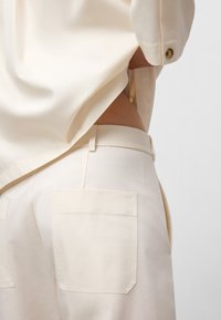Camicia avorio con maniche a risvolto, abbinata a pantaloni a vita alta con due grandi tasche quadrate e una texture liscia.