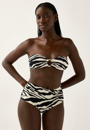 Nainen pukeutuneena mustavalkoiseen seepra-kuvioiseen bandeautyyppiseen bikiniin, jossa on keskellä kullanvärinen rengas, seisoo käsivarret vyötäröllä yksinkertaisen taustan edessä.