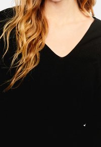 Rousse aux cheveux ondulés portant un haut noir à col V, montrée des épaules à la partie supérieure de la poitrine sur fond blanc.