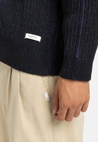 Maglione lavorato a maglia blu navy con texture a coste, dotato di una piccola etichetta Element Co. bianca; abbinato a pantaloni beige chiaro con pieghe.