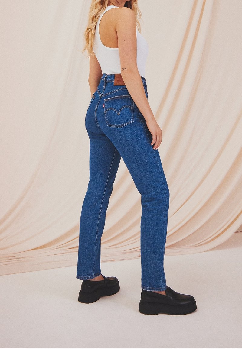 Femme portant un jean taille haute bleu, un haut sans manches blanc et des chaussures à plateforme noires, se tenant devant un fond en tissu beige.