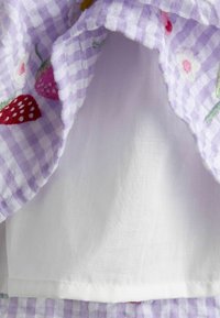 Tissu vichy lavande et blanc avec des fraises et des fleurs brodées, superposé sur un tissu en coton blanc uni en dessous.