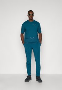 Το τιρκουάζ αθλητικό σετ περιλαμβάνει ένα κοντομάνικο μπλουζάκι με υφή και σχεδιαστική λεπτομέρεια, συνδυασμένο με ταιριαστά σκούρα παντελόνια και μαύρα υποδήματα.