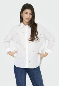 Camicia bianca con maniche corte, caratterizzata da ricami di ananas giallo e una vestibilità comoda, abbinata a jeans in denim blu.