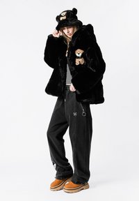 Chaqueta de piel sintética negra con bordado de osito de peluche, combinada con pantalones oscuros y holgados y botas marrones con plataforma. Incluye un gorro a juego con diseño de osito de peluche.