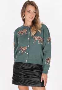 Cárdigan verde con patrones de tigre, que presenta un escote redondo, cierre de botones y botones decorativos. Combinado con una falda de cuero negra.
