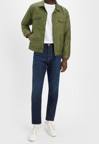 Grön nylonjacka med knäppning fram och två bröstfickor, kombinerad med mörkblå jeans och vita sneakers.