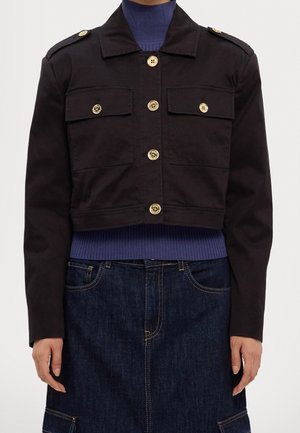 Sort cropped jakke med guldknapper, flap brystlommer, båret over en blå ribbet rullekrave og mørk denim nederdel.