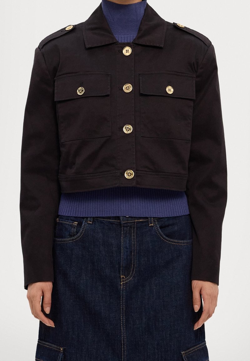 Veste courte noire avec boutons dorés, poches poitrine à rabat, portée sur un col roulé côtelé bleu et une jupe en jean foncé.