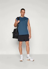 Sleeveless blue athletic shirt, black shorts, white sneakers with gum rubber soles, and a black shoulder bag. Model stands upright against a neutral background.