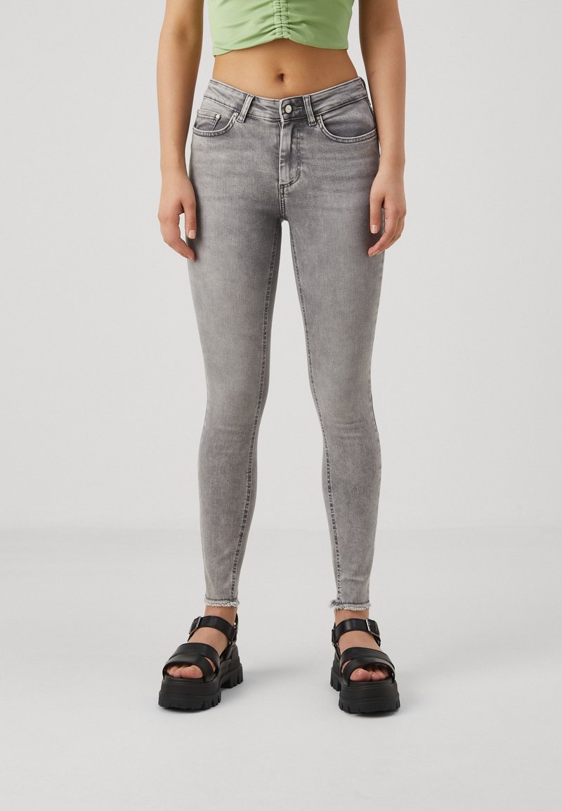 Personne portant un jean skinny gris clair, un haut court vert, et des sandales à plateforme noires, debout devant un fond uni.