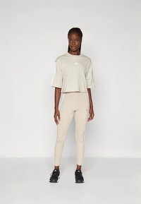 T-shirt bege cropped com logótipo branco da Adidas, combinado com leggings bege com detalhes brancos. Tênis desportivos pretos completam o conjunto.